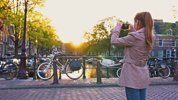 阿姆斯特丹度假的女性游客日落时站在运河上的桥上，用智能手机拍照