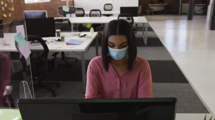 混血女商人戴着口罩坐在办公桌前使用电脑
