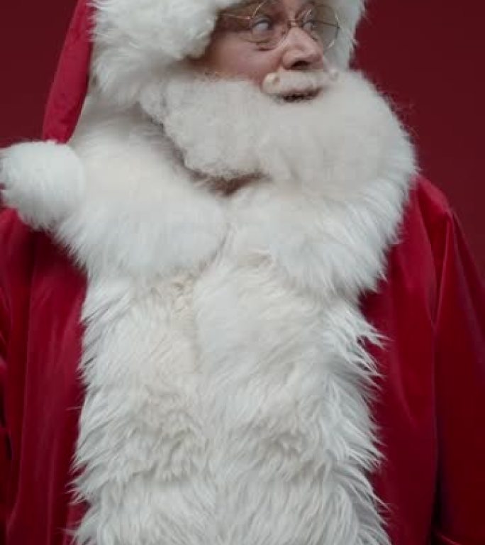 圣诞老人环顾四周并将手指放在嘴唇上的垂直视频，站在红色背景上