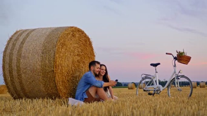 慢动作，一对成年夫妇在日落时坐在一吨重的草捆旁边看着电话的手持镜头