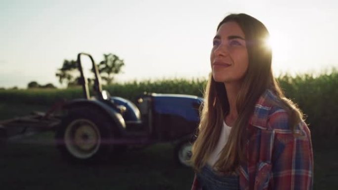 一个快乐的年轻女子的肖像看着背景为拖拉机的绿色玉米田地。女农场工人对自己的职业和农村生活方式感到满意