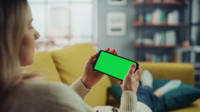 美丽的正宗女性躺在沙发上，在家里的客厅使用带有绿屏模拟显示的智能手机。她正在浏览互联网并在社交网络上