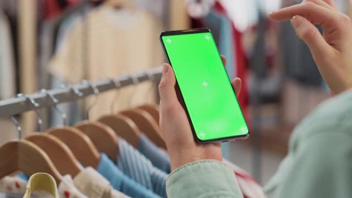 服装店: 女性使用色度键绿屏显示的智能手机。背景中带有时尚品牌商品的衣架，用于零售。特写移动设备的镜
