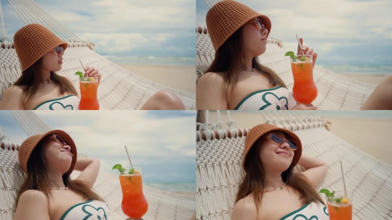 快乐的年轻女子，喝鸡尾酒，在海滩的吊床上放松。