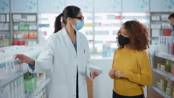 新型冠状病毒肺炎药房戴口罩: 专业药剂师帮助美丽的女性顾客推荐药物，交谈。药店里到处都是药物，药丸，