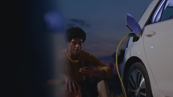SLO MO肖像，一名年轻的篮球运动员使用智能手机，晚上在停车场给电动车充电