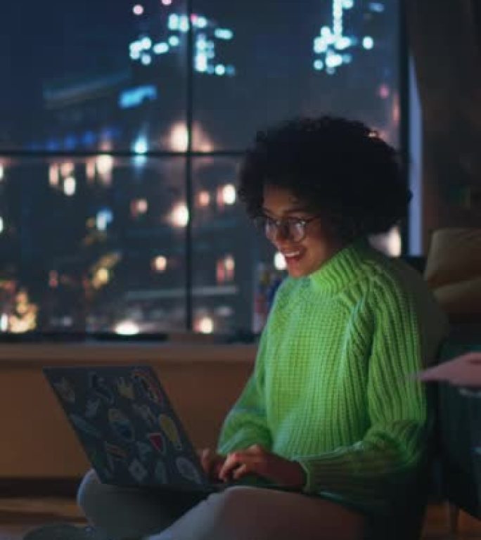 积极的拉丁女性在家里使用带贴纸的笔记本电脑。女人躺在地板上，微笑着在电脑上发短信。窗户后面的夜景。垂