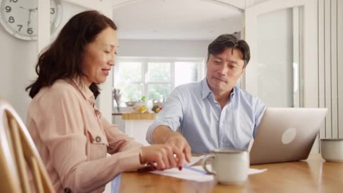 成熟的亚洲夫妇坐在家里的餐桌旁，用笔记本电脑整理家庭账单和财务 -- 慢动作拍摄