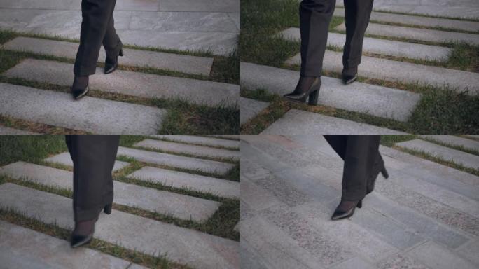 穿着高跟鞋和经典裤子的女性脚的特写镜头优雅地登上混凝土楼梯