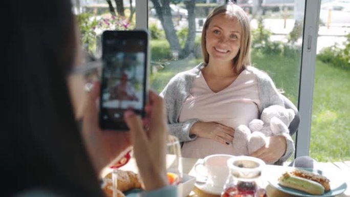 快乐孕妇与泰迪熊一起为智能手机相机摆姿势，而朋友在咖啡馆拍照