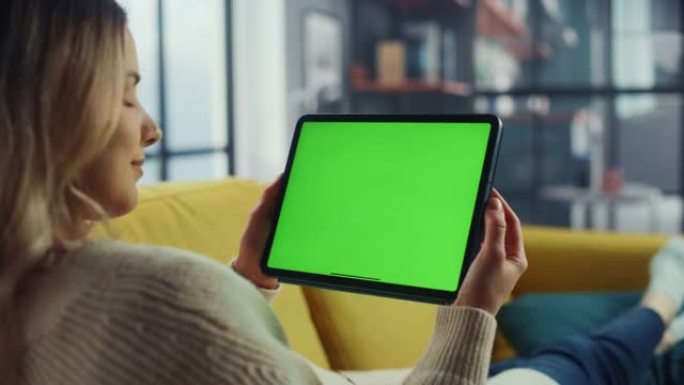 美丽的正宗女性使用平板电脑与绿屏模拟显示在家里的客厅，而躺在沙发上。她正在浏览互联网并在社交网络上查