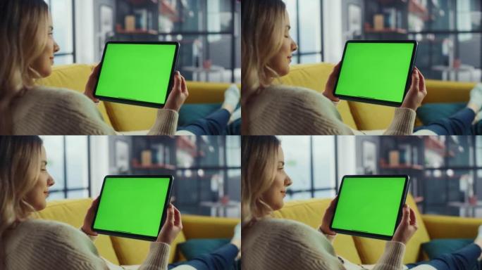 美丽的正宗女性使用平板电脑与绿屏模拟显示在家里的客厅，而躺在沙发上。她正在浏览互联网并在社交网络上查