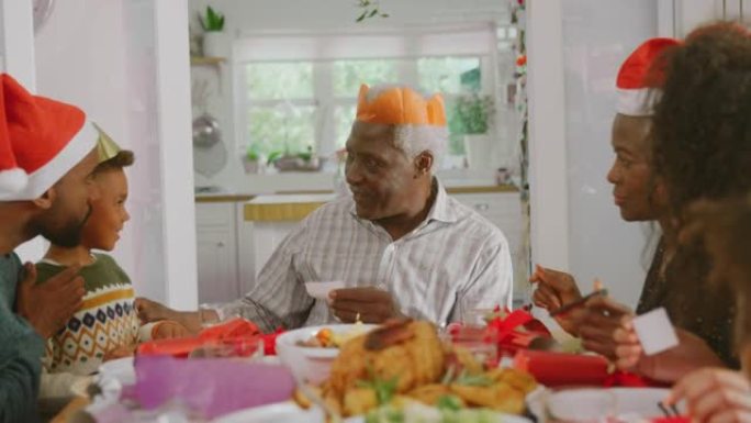 戴纸帽的多代家庭在一起吃饭的同时阅读圣诞饼干笑话