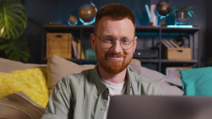年轻快乐聪明的白人男性自由职业者的肖像在眼镜上使用笔记本电脑在家工作，看着相机。