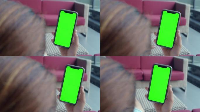 使用带有绿色模拟屏幕的智能手机，在酒店大堂坐在沙发上的年轻女子的肩膀拍摄，特写镜头