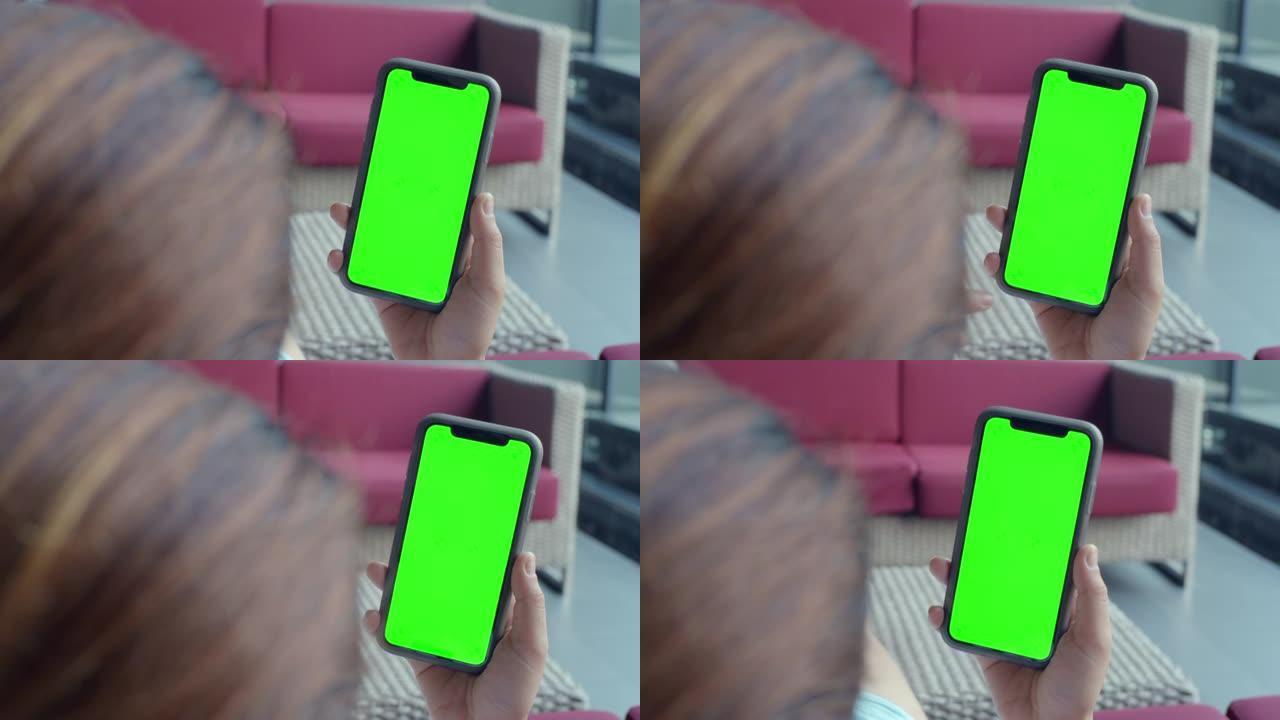 使用带有绿色模拟屏幕的智能手机，在酒店大堂坐在沙发上的年轻女子的肩膀拍摄，特写镜头
