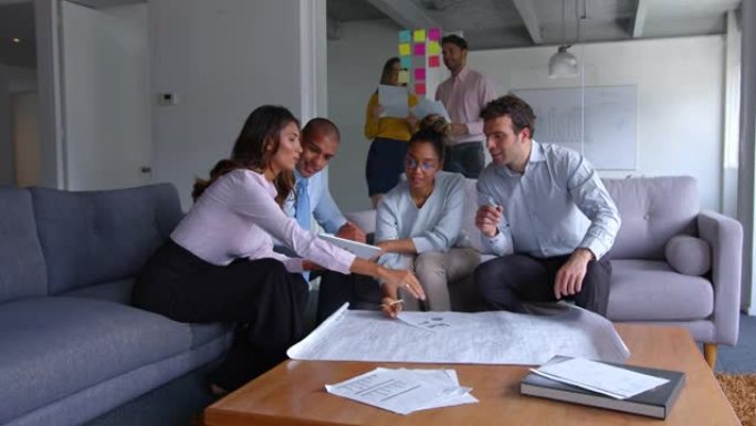 办公室休息室的一群建筑师坐在沙发上看着蓝图，同时讨论项目的所有想法