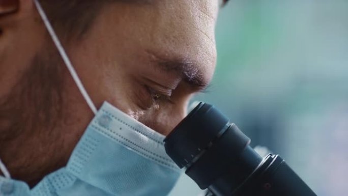 宏观特写一位英俊的男性科学家戴着医用口罩，看着显微镜的镜头。微生物学家使用技术设备在现代实验室中研究