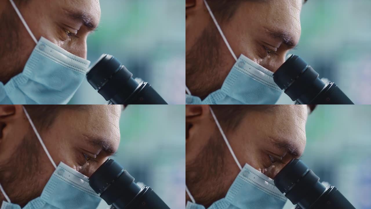 宏观特写一位英俊的男性科学家戴着医用口罩，看着显微镜的镜头。微生物学家使用技术设备在现代实验室中研究