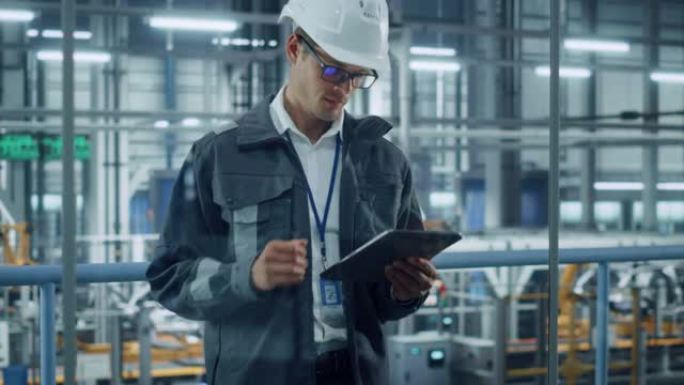 穿着制服和安全帽的英俊工程师在汽车装配厂使用平板电脑。从事车辆设计的工业专家，俯瞰技术设施的生产。
