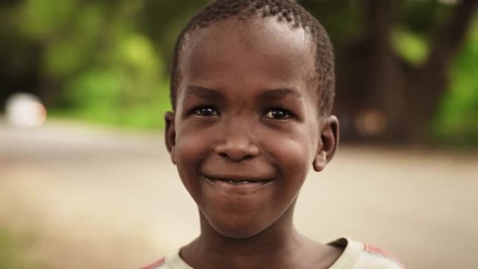 一个害羞的真实的非洲男孩在镜头前微笑，背景是一个模糊的乡村。黑人男孩代表未来、希望和接纳。纪录片镜头