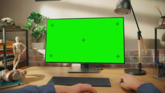 在具有绿屏模拟显示的台式计算机上工作的人的POV第一人称视图。男性坐在办公桌前，在时尚的客厅里浏览互