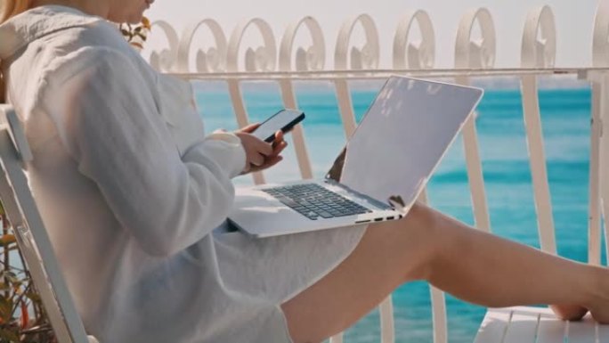 金发碧眼的女人在旅馆房间的阳台上工作时检查她的智能手机，膝上放着笔记本电脑，背景是大海