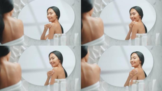 美丽的亚洲女人将润肤霜涂在她完美的肩膀上，使皮肤柔软，在镜子中性感地微笑。快乐的女性享受她的美丽。健