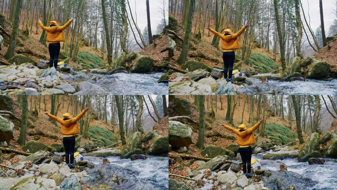 伸出双臂的女人站在森林里流水旁的岩石上