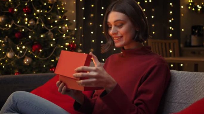 一个穿着红色毛衣的年轻漂亮女人微笑着打开一个红色的礼品盒，里面温暖的光线照亮了她的脸，然后高兴地关上