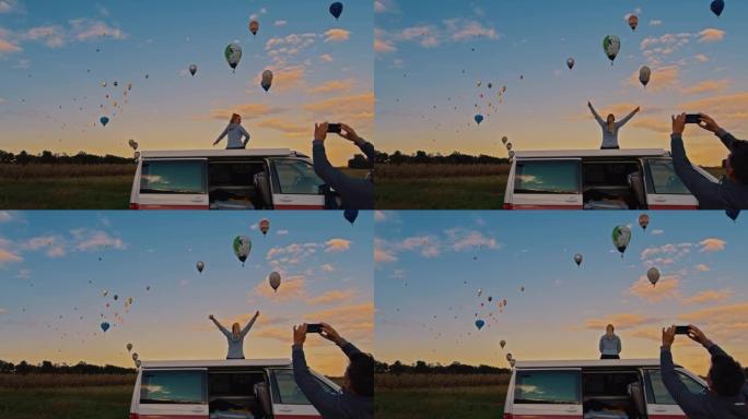 带着照相手机的情侣看着热气球飞过田野