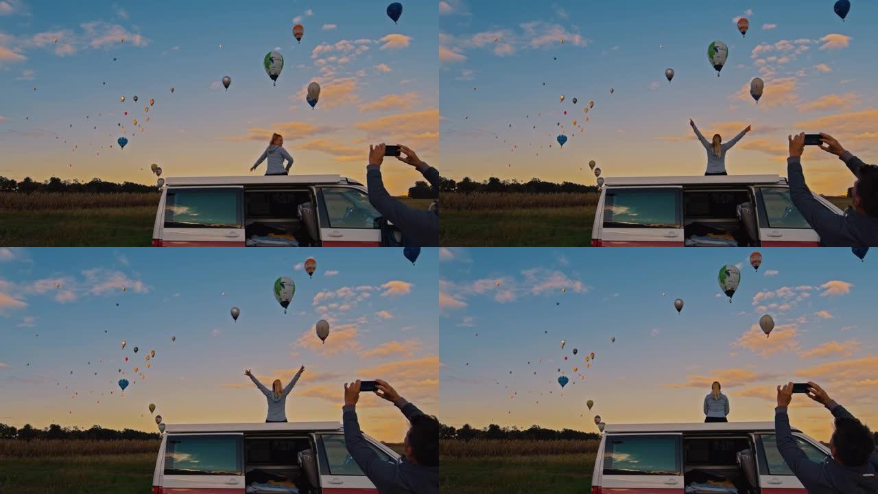带着照相手机的情侣看着热气球飞过田野
