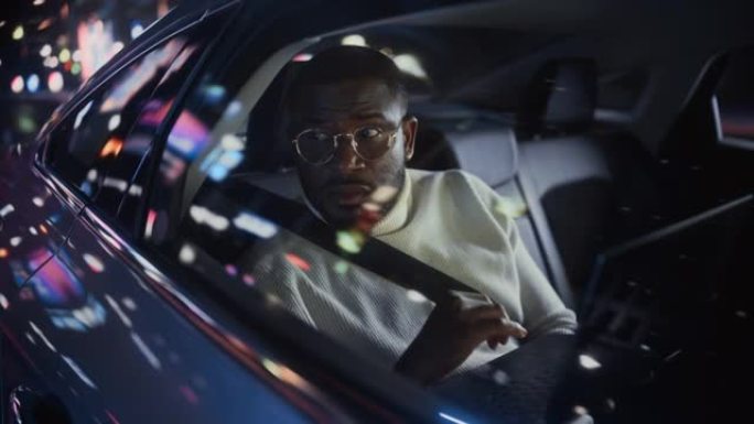 时髦的戴眼镜的黑人在晚上坐出租车的后座上下班。男性使用笔记本电脑，在城市街道上开着霓虹灯的汽车时看着