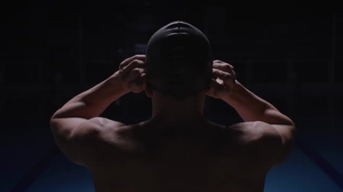 年轻的职业男游泳运动员的电影后视图镜头正在戴上泳帽，然后开始在游泳池里努力练习和训练。运动、竞争、胜
