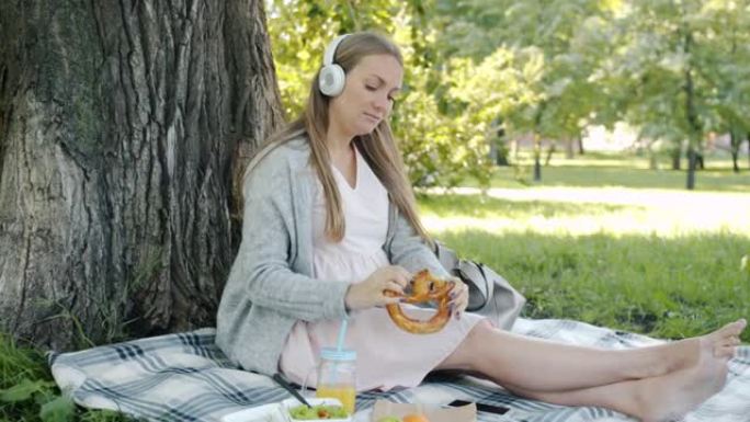 快乐的孕妇戴着耳机听音乐，在公园户外野餐