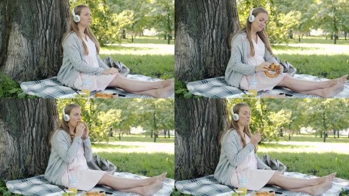 快乐的孕妇戴着耳机听音乐，在公园户外野餐