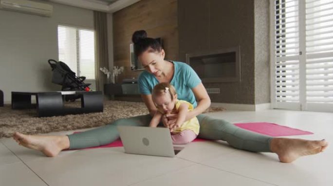 白人母亲抱着婴儿在家里笔记本电脑前的瑜伽垫上练习瑜伽