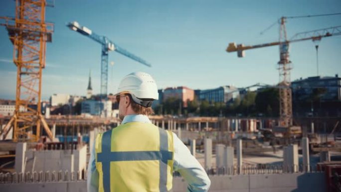充满自信的大胡子的土木工程师-戴着墨镜的建筑师站在外面，背对着镜头在一个建筑工地上。男人戴着安全帽，