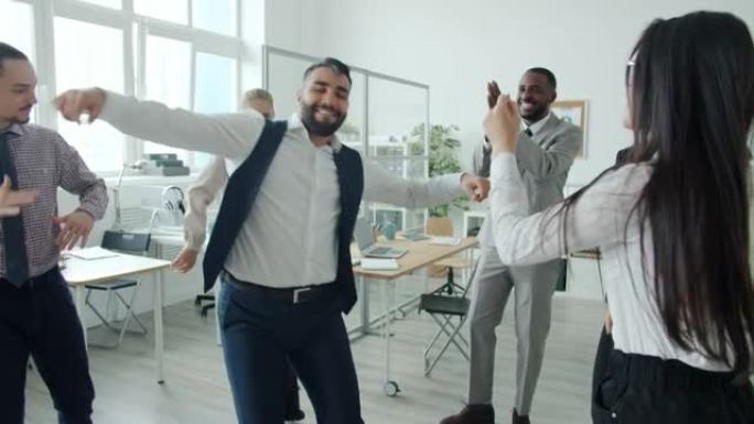 穿着正式服装的男人的慢动作在办公室跳舞，而同事们却在玩乐和鼓掌