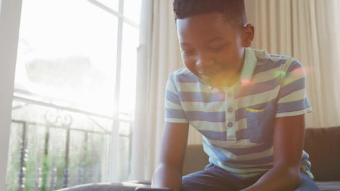 快乐的非洲裔美国男孩坐在沙发上，用智能手机微笑，被阳光照射