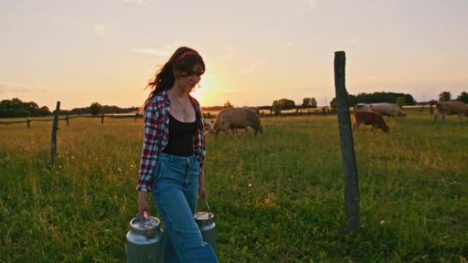 SLO MO Young女牧场主在日落时沿着牧场携带两个牛奶罐