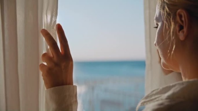 金发女人从酒店房间的窗户向外望去，手里拿着透明的窗帘，背景是大海的阳台