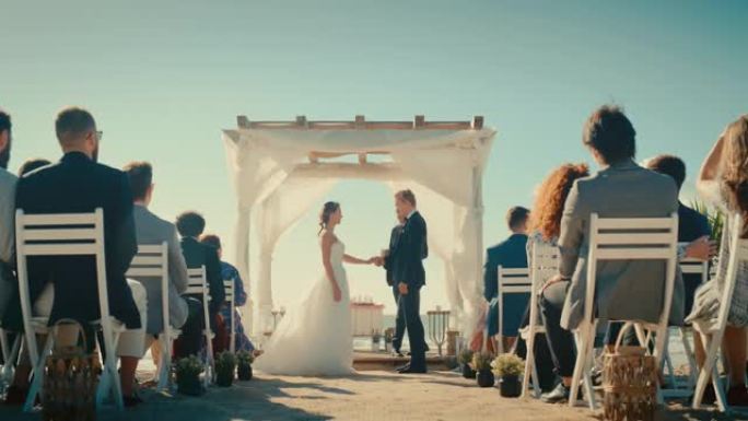 美丽的新娘和新郎在海边的户外婚礼上。浪漫情侣结婚、交换戒指、亲吻和与多民族朋友分享庆祝活动的完美场所
