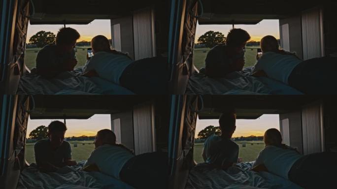 WS夫妇使用智能手机从日落时停在草地上的露营车的小屋里拍照