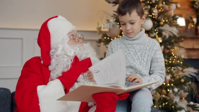 可爱的男孩和圣诞老人在元旦在装饰好的树附近聊天和看书