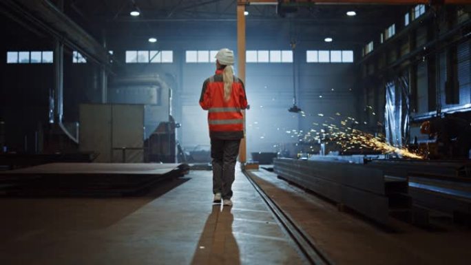 专业重工业工程师/穿着安全制服和安全帽的工人使用平板电脑。严肃成功的女性工业专家走在金属制造仓库里。