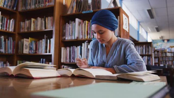 戴着蓝色头巾的亚洲女学生坐在打开书本的书桌前做笔记