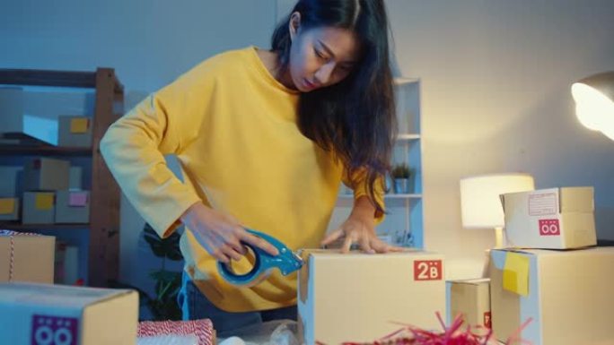 年轻的亚洲女商人准备产品使用胶带包装盒，以便在晚上发送到家庭办公室的客户采购订单。