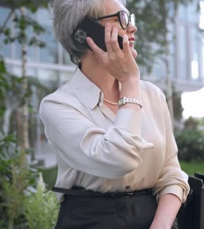 一位戴着时尚眼镜的已婚时尚中年女商人在电话中愉快地交谈，而相机则将她从一侧拍摄到另一侧