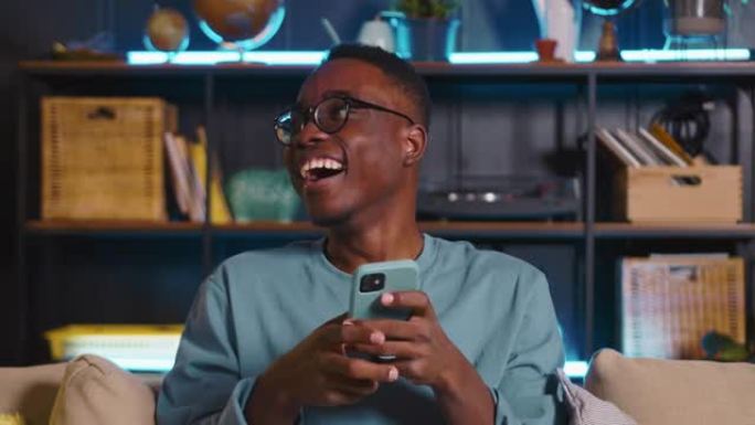 年轻快乐的黑人男学生笑着，在舒适的家中在线社交网络上观看有趣的模因视频。